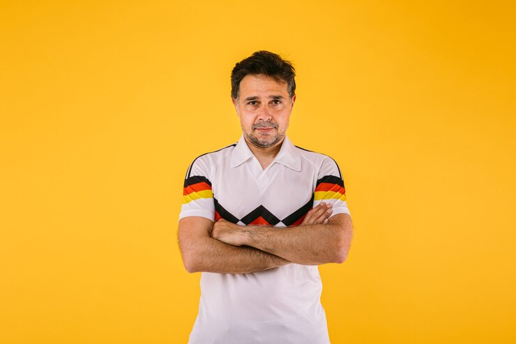 Foto fã de futebol vestindo uma camiseta branca com listras pretas vermelhas e amarelas posa com os braços cruzados