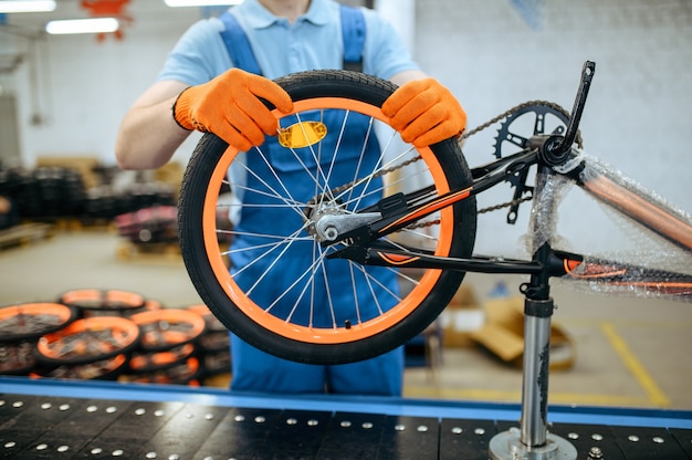 Fábrica de bicicletas, trabajador en línea de montaje, instalación de ruedas. Macho mecánico en uniforme instala piezas de ciclo en taller