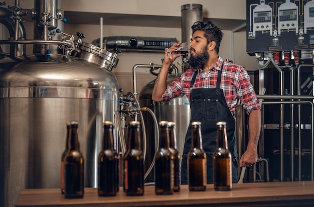 Fabricante masculino hipster tatuado barbudo indio degustando y presentando cerveza artesanal en la microcervecería.