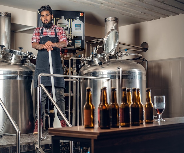 Fabricante masculino hipster tatuado barbudo indio que presenta cerveza artesanal en la microcervecería.