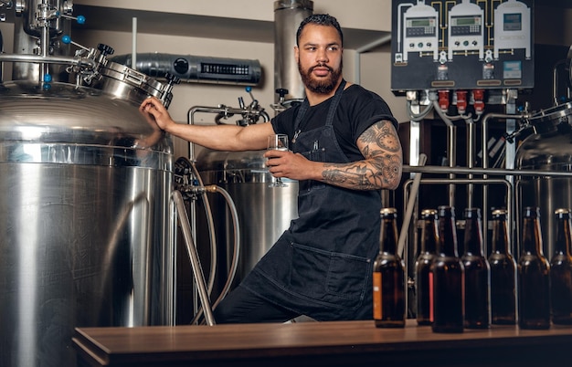 Fabricante masculino hipster tatuado de barba preta, degustação de cerveja na microcervejaria.