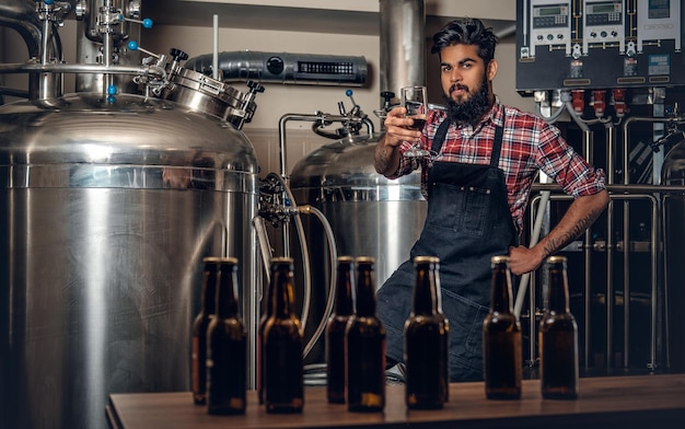 Fabricante masculino hipster tatuado e barbudo indiano degustando cerveja artesanal na microcervejaria.