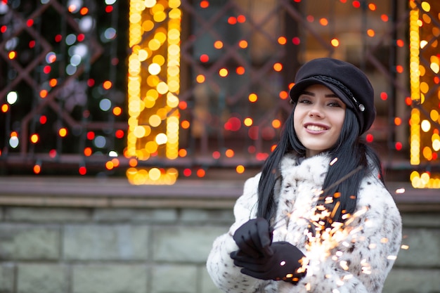 Fabulosa mujer morena con ropa de abrigo celebrando las vacaciones de invierno con bengalas en la calle. Espacio para texto