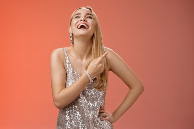 Fabulosa mujer rubia atractiva despreocupada en vestido de fiesta de noche plateado riendo a carcajadas diviértete levanta la mano con alegría apuntando el dedo índice derecho disfrutando de un increíble sentido del humor, fondo rojo.