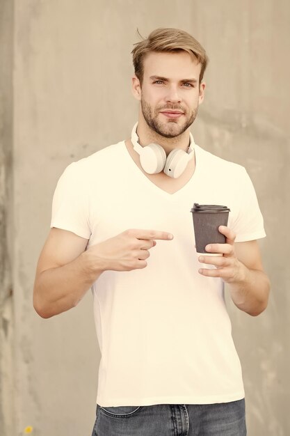 Foto faça seu pedido de café goto homem bonito aponte para xícara de café ao ar livre cara jovem bebe café de manhã nova tecnologia vida moderna desfrutando de café e música