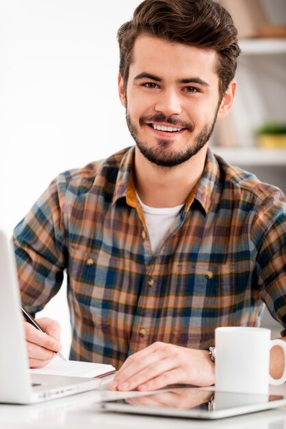 Face à satisfação no trabalho. Jovem sorridente escrevendo em um bloco de notas e olhando para a câmera enquanto está sentado em seu local de trabalho