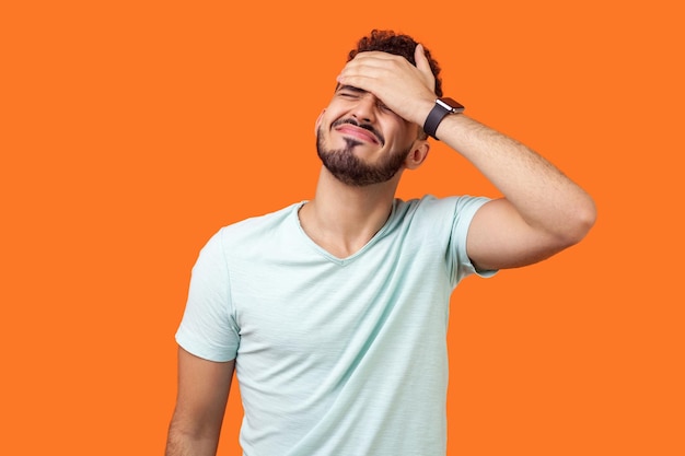 Facepalm Retrato de un hombre moreno desesperado con barba en camiseta blanca cubriendo la cara con la mano sintiéndose arrepentido y culpándose a sí mismo por el error en el estudio interior aislado en fondo naranja