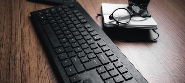 Fahnennahaufnahme des hölzernen Schreibtisches mit Tastatur, Gläsern und Notizbuch. Von zuhause aus arbeiten. Technologie-Tastatur