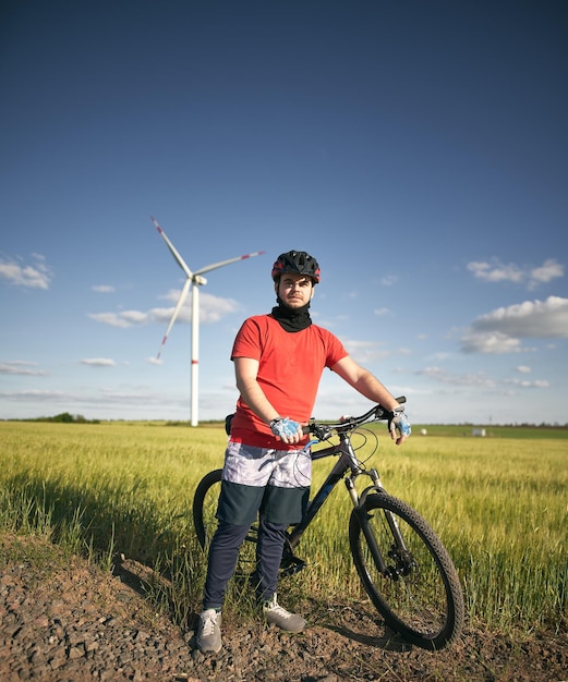 Fahrradfahrer mit fahrradhelm, der während des sommerabends vor der windkraftanlage mit dem mountainbike steht