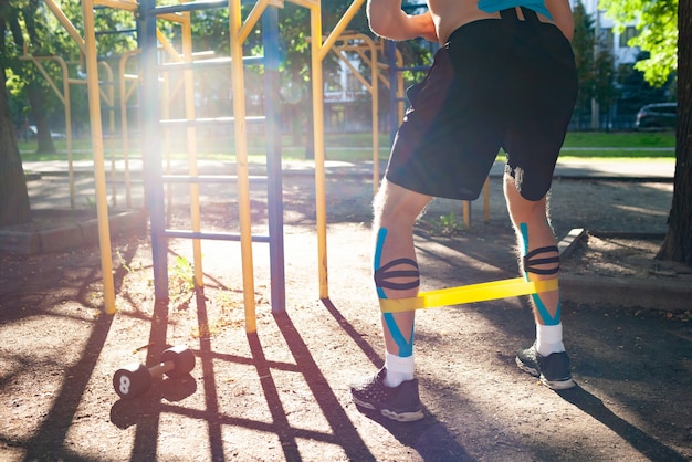 Faixa de resistência de aptidão de treinamento de homem musculoso nas pernas no campo de esportes. Vista traseira do jovem fisiculturista irreconhecível com cinesiologia elástica, gravando no treinamento corporal ao ar livre. Conceito de reabilitação.