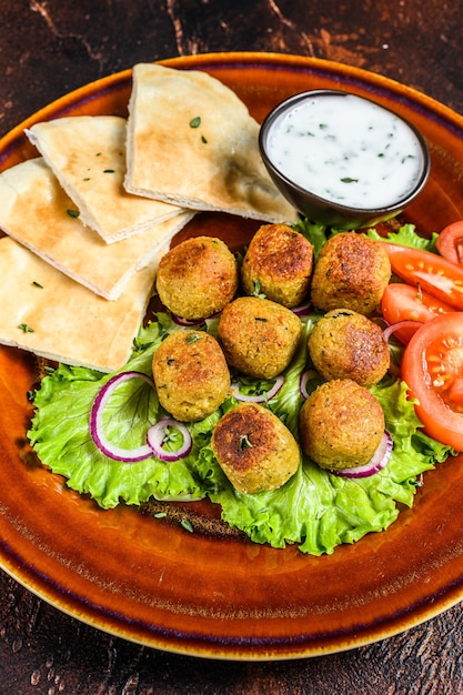 Falafel vegetariano com pão pita, legumes frescos e molho no prato. Fundo escuro. Vista do topo.