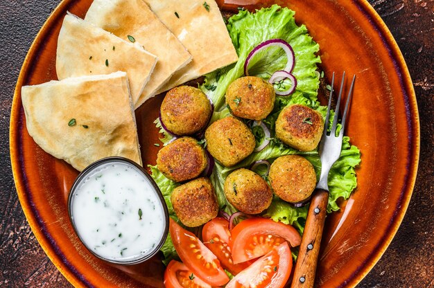 Falafel vegetariano com pão pita, legumes frescos e molho no prato. Fundo escuro. Vista do topo.