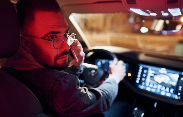 Falando por telefone Iluminação vermelha Bonito homem sem barba em roupas da moda andando de automóvel