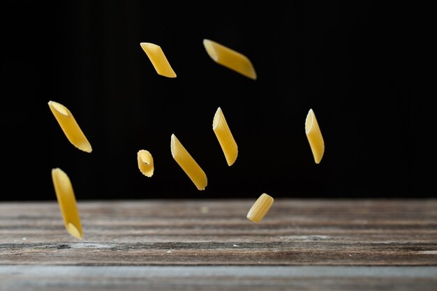 Fallende Penne-Nudeln. Fliegende gelbe rohe Makkaroni über schwarzem Hintergrund und Rückprall vom Holztisch. Flacher DOF.