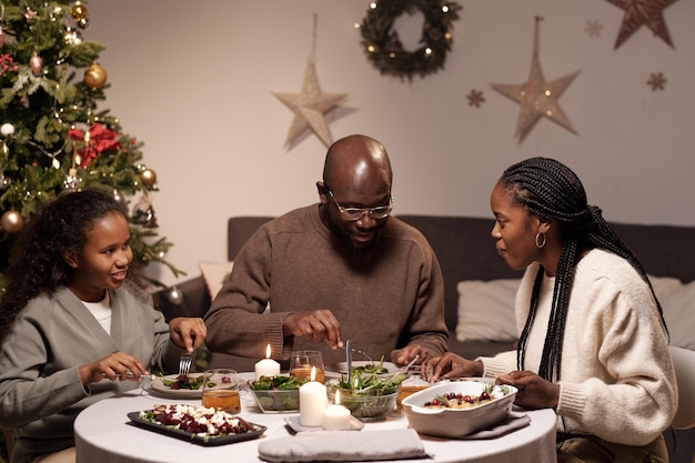 Família africana feliz de três pessoas interagindo em uma mesa festiva servida enquanto comia comida caseira tradicional de natal