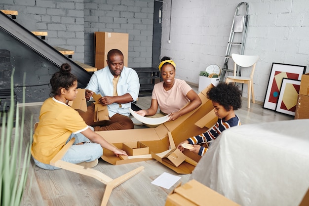 Familia africana joven de sentarse en el suelo de la gran sala de estar mientras desembala y saca las piezas de los muebles de la caja de cartón