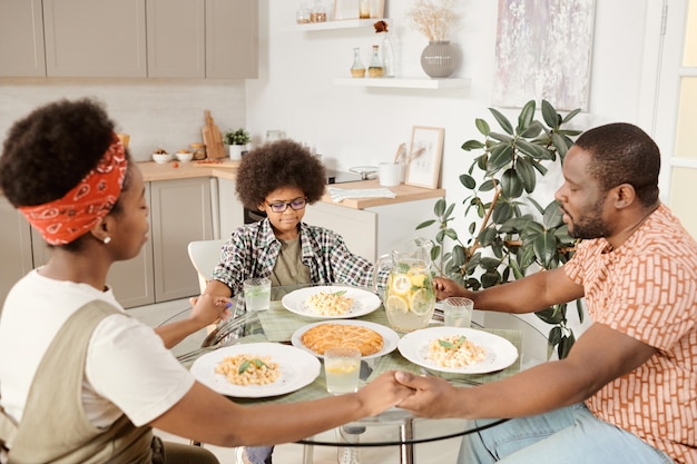 Familia africana joven de tres orando antes de la cena con los ojos cerrados