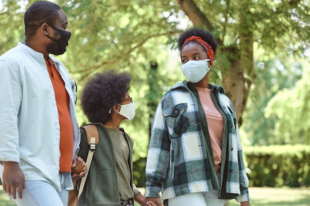 Familia africana de tres en máscaras protectoras caminando en el parque el día de verano