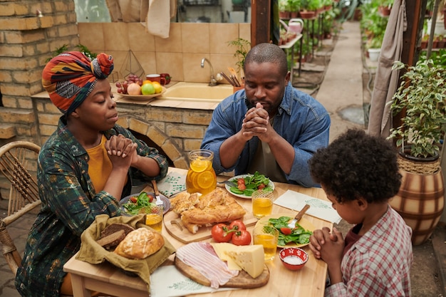 Familia africana de tres sentados en la mesa del comedor y orando juntos antes de la cena