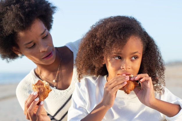 Família afro-americana no piquenique na praia. Mãe e filha em roupas casuais sentadas no cobertor, comendo pães. Família, relaxamento, conceito de natureza