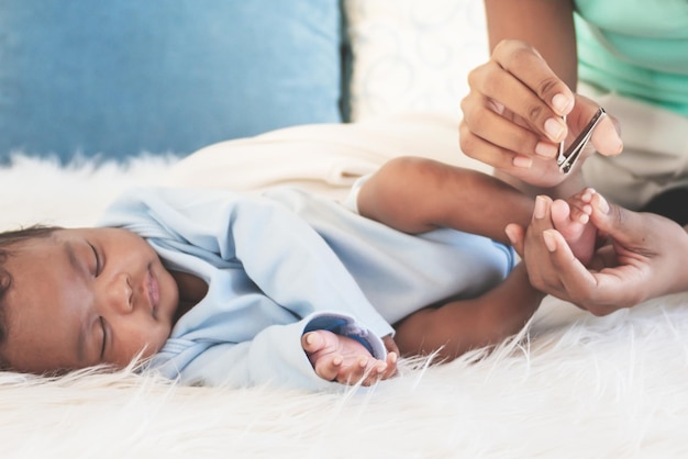 Familia afroamericana Una madre está usando un cortador de uñas para su hijo recién nacido de 1 mes que está durmiendo en una cama blanca para el cuidado de la salud y el concepto de bebé recién nacido