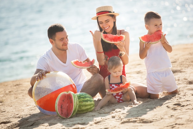 Familia alegre comiendo sandía en la playa. Los niños pequeños y sus padres en la orilla del mar se divierten. Familia alegre en la orilla del mar