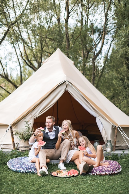 Familia alegre comiendo sandía sentado al aire libre cerca de la casa Bigi Tipi en el parque