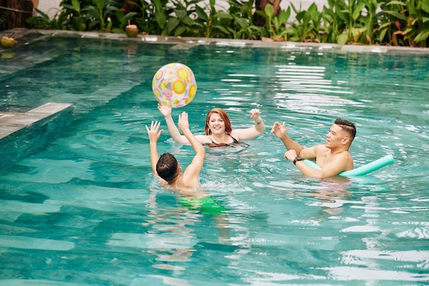 Família alegre de três pessoas jogando bola inflável um para o outro ao brincar na piscina do spa resort