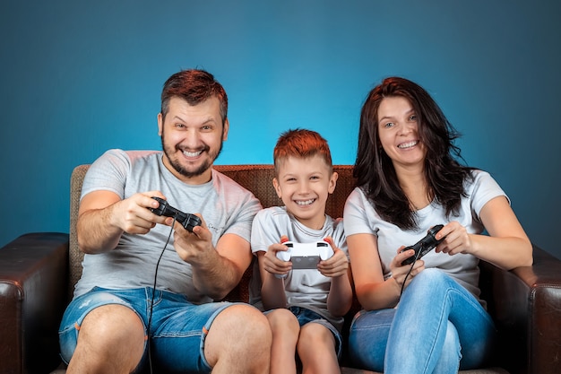 Una familia alegre, padre, madre e hijo juegan en la consola, los videojuegos, reaccionan emocionalmente sentados en el sofá. Día libre, entretenimiento, ocio, pasar tiempo juntos.