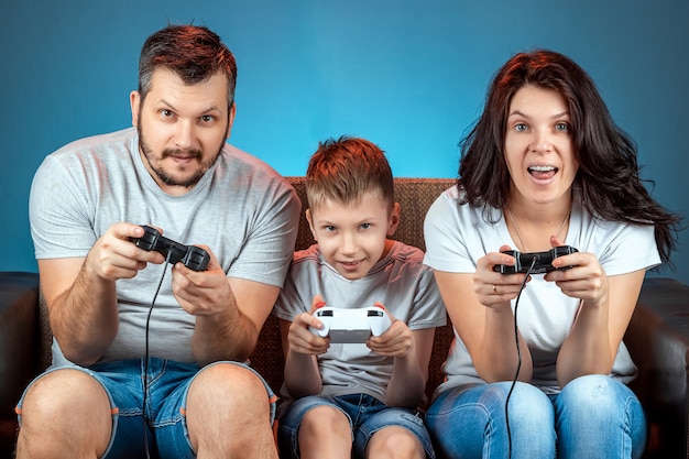 Una familia alegre, padre, madre e hijo juegan en la consola, los videojuegos, reaccionan emocionalmente sentados en el sofá. Día libre, entretenimiento, ocio, pasar tiempo juntos.