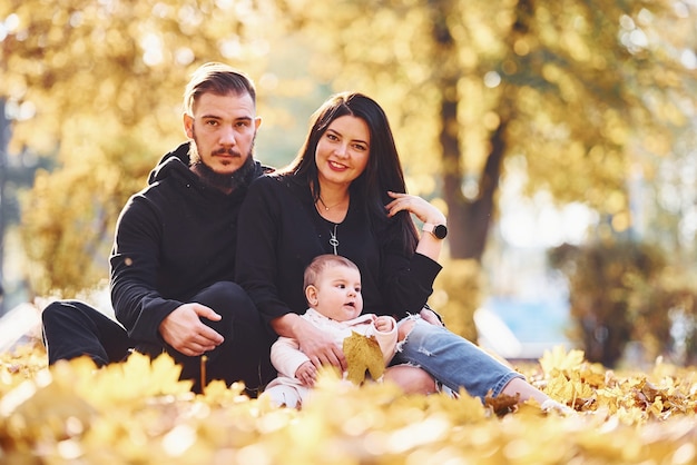 La familia alegre se sienta en el suelo y se divierte junto con su hijo en el hermoso parque otoñal.