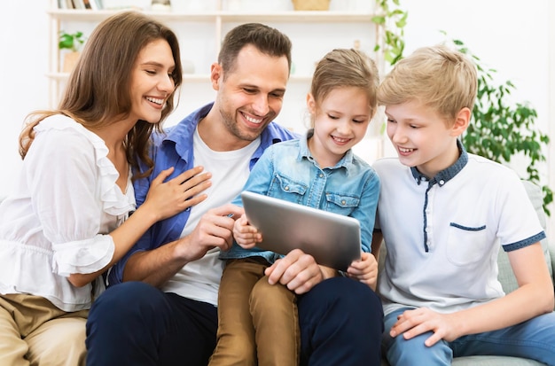 Família alegre usando computador tablet passar tempo juntos sentados no sofá dentro de casa e assistindo desenhos animados. Fim de semana em casa.