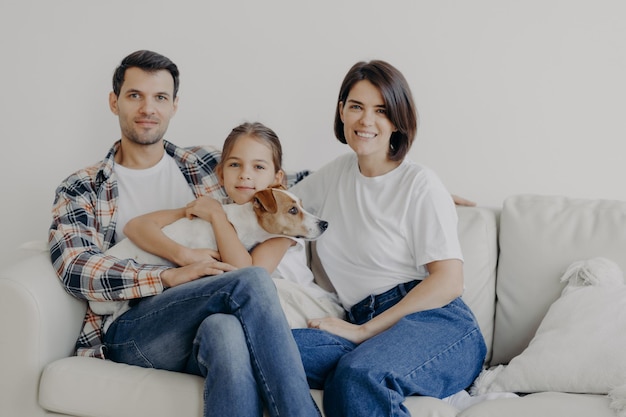 Família amigável posar juntos no sofá desfruta de atmosfera doméstica Pai mãe sua filhinha e cachorro com pedigree passam o fim de semana em casa posam na sala de estar têm expressões de rosto feliz