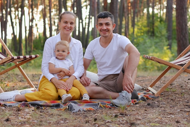 Família amorosa com menina infantil fofa sentada na floresta no chão na manta olhando para a câmera expressando emoções positivas e felicidade curtindo a bela natureza e passatempo juntos