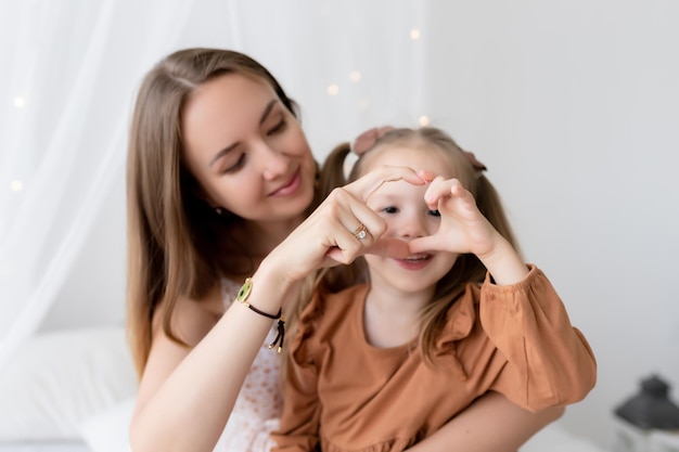 Família amorosa feliz Mãe e filha brincam e se divertem na cama no quarto Eles mostram um coração com as mãos O prazer de ser mãe parabéns pelo dia das mães