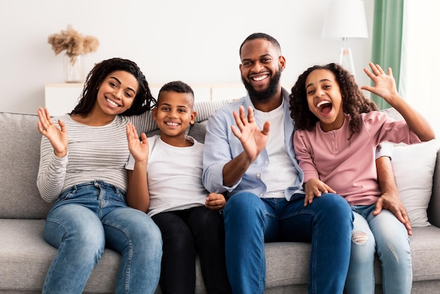Família amorosa feliz. Retrato de alegre homem, mulher, menino e menina afro-americanos sentados no sofá em casa, posando para foto e acenando com as mãos para a câmera. Quatro jovens negros sorridentes