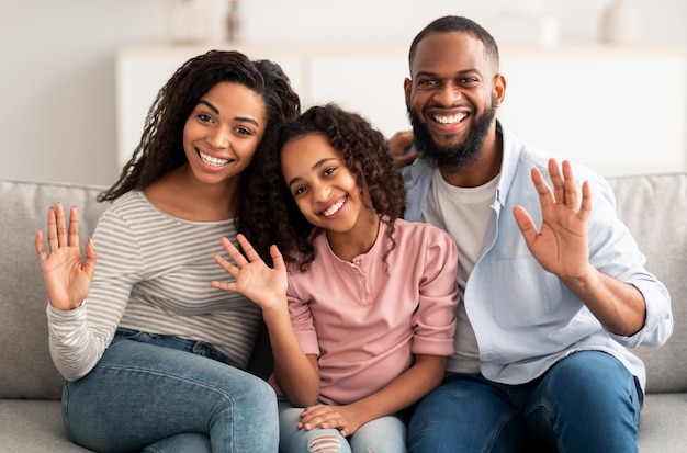 Família Amorosa Feliz. Retrato de um homem afro-americano alegre, mulher e menina sentada no sofá em casa, posando para a foto e acenando com as mãos para a câmera. Jovens negros sorridentes