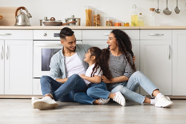 Família árabe feliz, passando o fim de semana juntos em casa, alegre jovem pai do Oriente Médio, mãe e filhinha linda sentada no chão de ladrilhos da cozinha branca, conversando, copie o espaço