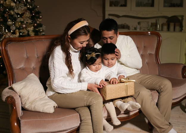 Familia asiática con dos niños abre regalos mientras está sentado en el sofá junto al árbol por la noche en casa