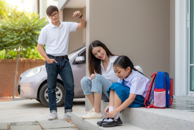 Familia asiática con padre, madre mirando a su hija estudiantes de preescolar en uniforme a usar sus propios zapatos