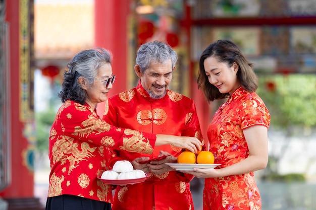 Una familia asiática con padres mayores en vestido rojo cheongsam qipao está ofreciendo comida al dios ancestral dentro del templo budista chino durante el año nuevo lunar para los mejores deseos de bendición y buena suerte