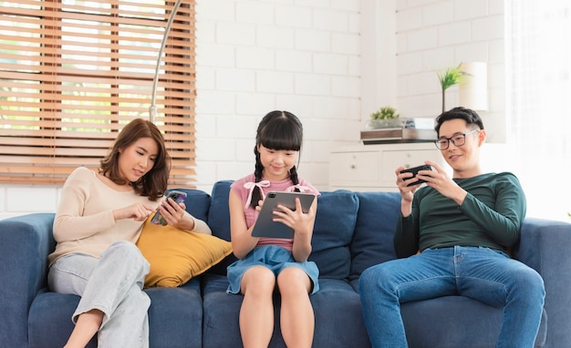 Família asiática relaxa no sofá usando tecnologia digital de gadgets de telefones inteligentes.