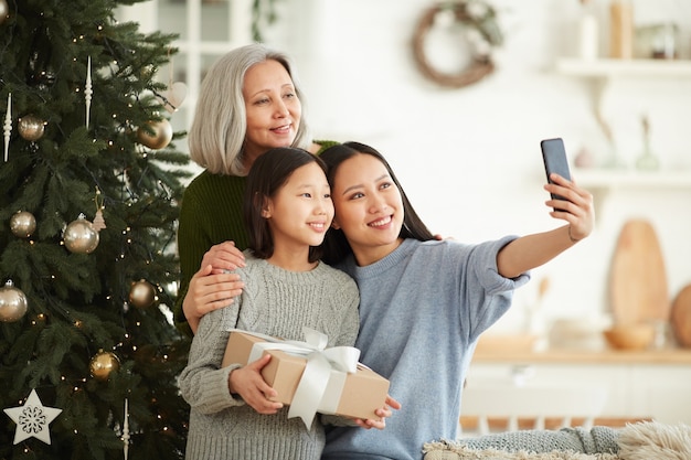 Familia asiática de tres haciendo retrato selfie en teléfono móvil de pie cerca del árbol de Navidad durante las vacaciones