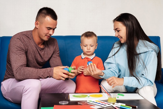 La familia blanca feliz se dedica al trabajo creativo y se divierte en casa. Mamá, papá e hijo pequeño pintan y esculpen con plastilina