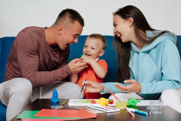 La familia blanca feliz se dedica al trabajo creativo y se divierte en casa. Mamá, papá e hijo pequeño pintan y esculpen con plastilina