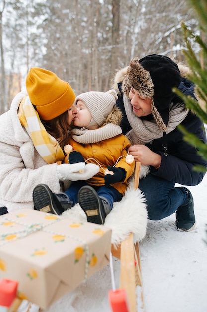 Familia en el bosque de invierno, padres junto al bebé en un trineo, mamá besa al bebé