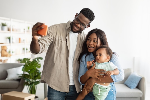 Familia cariñosa feliz. Retrato de amorosa familia negra tomando selfie con su hijo de pie en la sala de estar en casa. Hombre que usa el teléfono inteligente para la foto, celebrando el día de la mudanza en la nueva casa, capturando el momento