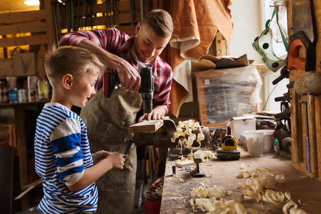 família, carpintaria, marcenaria e conceito de pessoas - pai e filho pequeno com broca perfurando prancha de madeira na oficina