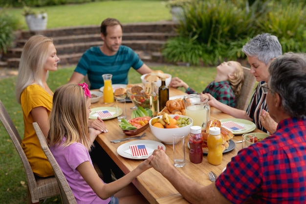 Família caucasiana de três gerações de mãos dadas dizendo graça antes de comer a refeição juntos no jardim. família de três gerações comemorando o dia da independência comendo ao ar livre juntos.