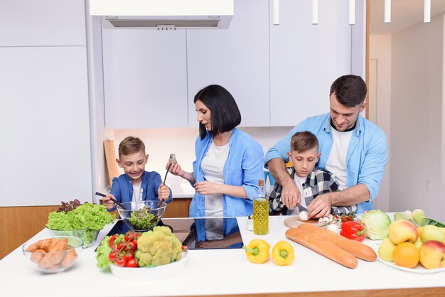 Família jovem e elegante com dois filhos preparando um café da manhã  vegetariano saudável com legumes frescos na cozinha